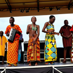 **Vibrant Rwandan Cultural Fusion:** A dynamic and energetic scene showcasing the modern face of Rwandan culture. A charismatic young Rwandan musician, similar to Meddy, is performing live on stage, embodying a blend of R&B and Afrobeat with Kinyarwanda lyrics. Behind them, band members play a mix of traditional Rwandan instruments like the Inanga and Umuduri, alongside contemporary electronic synthesizers. Interspersed around the stage or in the audience, models are stylishly dressed in vibrant fashion pieces crafted from traditional Kitenge fabrics, reinterpreted with modern, sleek designs that reflect global trends. The atmosphere is filled with bright colors, pulsating rhythms, and a sense of youthful optimism and global appeal, perhaps set against a backdrop suggesting a bustling Kigali street or a modern concert venue.